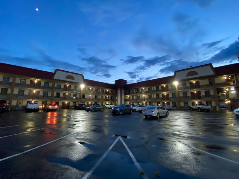 A three-story hotel with exterior corridors is shown at dusk. Several cars are parked in the wet parking lot, reflecting the light from the building and the dark, cloudy sky above. The central entrance is illuminated by outdoor lights.