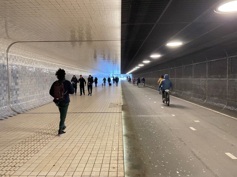 A long tunnel with white tiled walls and a high ceiling is divided into two lanes. The left lane has pedestrians walking, and the right lane has cyclists. The tunnel is well-lit with round ceiling lights, and people are moving towards the bright exit.