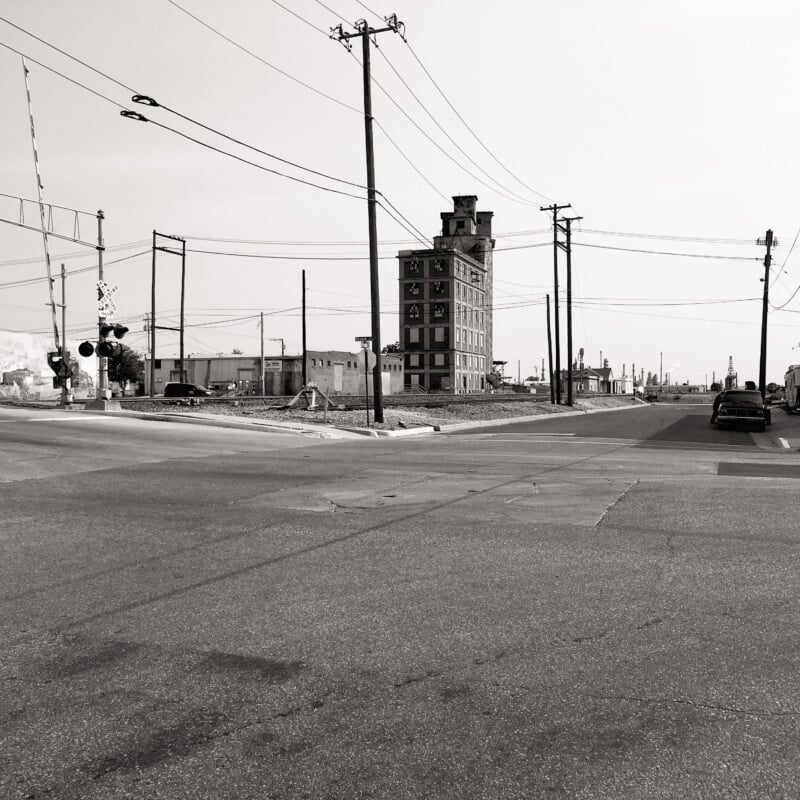 Black-and-white photo of an urban street with intersecting power lines, traffic lights, and a tall, narrow, derelict building in the center. Nearby, there are industrial structures and a car parked on the right side of the road. The overall scene looks deserted.