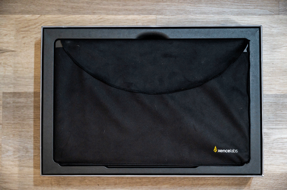 A black protective sleeve with the Xencelabs logo in the bottom right corner is positioned inside an open dark-colored box, which is placed on a wooden surface. The sleeve appears to be designed for a tablet or laptop.