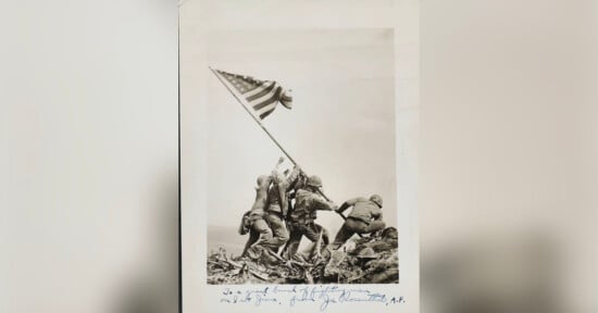 A historic black-and-white photograph depicting six soldiers raising the American flag on Iwo Jima during World War II. The photo shows the soldiers working together to plant the flagpole in the ground amid rugged terrain. Handwritten text is visible at the bottom.