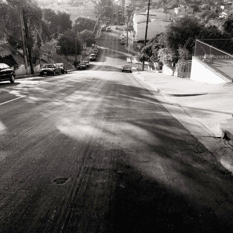 A black-and-white photograph of a steep city street with parked cars on either side and houses lining the road. The sky is bright, casting shadows from trees and buildings. The street extends uphill, leading towards more urban structures in the distance.