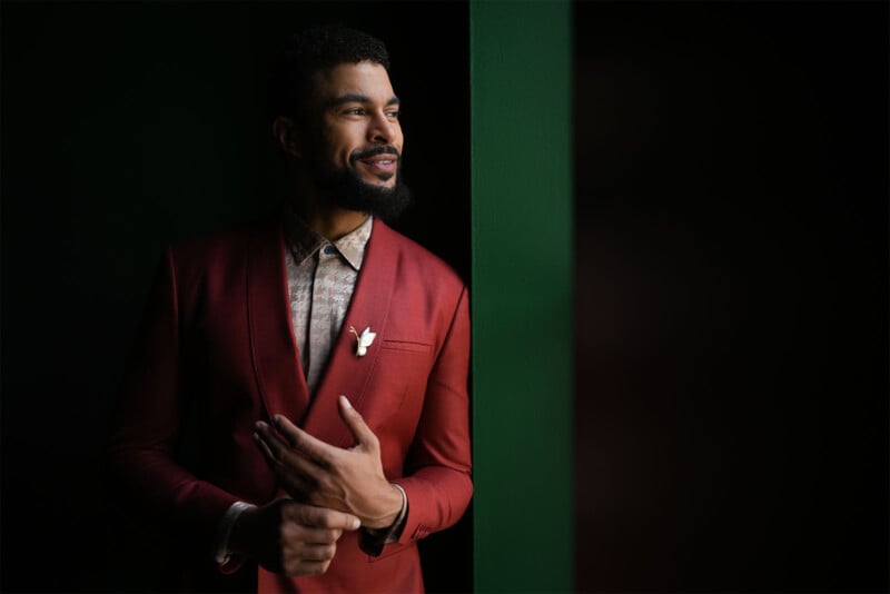 A man with a beard and short curly hair is standing next to a green wall. He is wearing a red suit jacket over a patterned shirt and has a small white flower pinned to his lapel. He smiles while looking off to the side, illuminated by soft lighting.