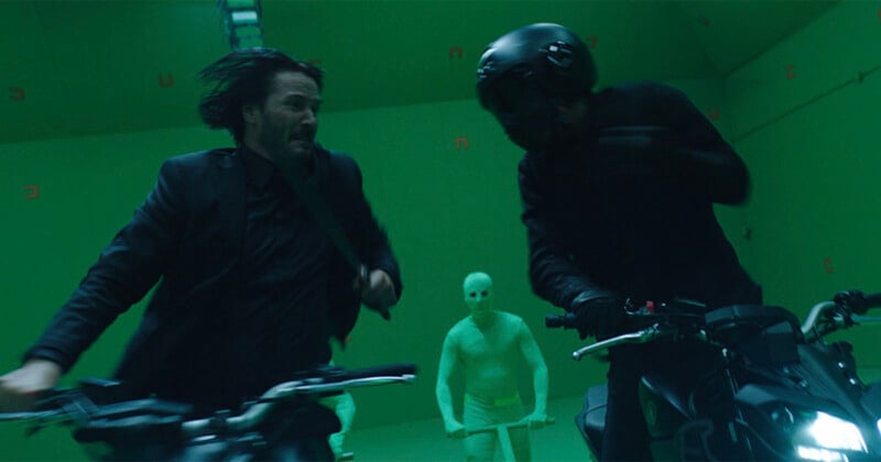 A scene featuring two motorcyclists, one with shoulder-length black hair and the other wearing a helmet, engaged in an intense chase. The background is green, resembling a studio setup with a person in a full-body green suit holding a prop.