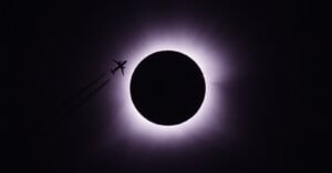 A plane is silhouetted against the dramatic backdrop of a solar eclipse, with the moon almost completely covering the sun, leaving only a bright halo of light. The sky around is dark, highlighting the striking contrast between the airplane and the eclipse.