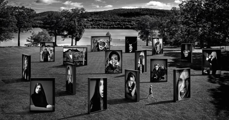 Black and white photo of a park with numerous large, vertical frames displaying intense, expressive portraits of people. In the background, there are trees and a body of water. Two individuals walk among the frames.