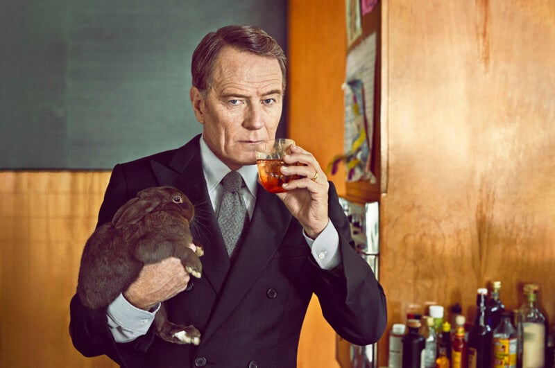 A man in a suit holds a drink in one hand and a bunny in the other. He is standing indoors near a wall with various items and a shelf with bottles. The man has a serious expression as he gazes at the camera.