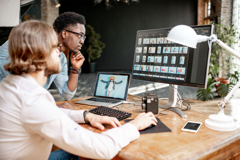 Two men work together at a wooden desk in a modern office. One man, wearing glasses, types on a keyboard connected to a computer with photo thumbnails on the screen. The other man, also wearing glasses, intently observes the screen. A camera, lamp, and phone are on the desk.