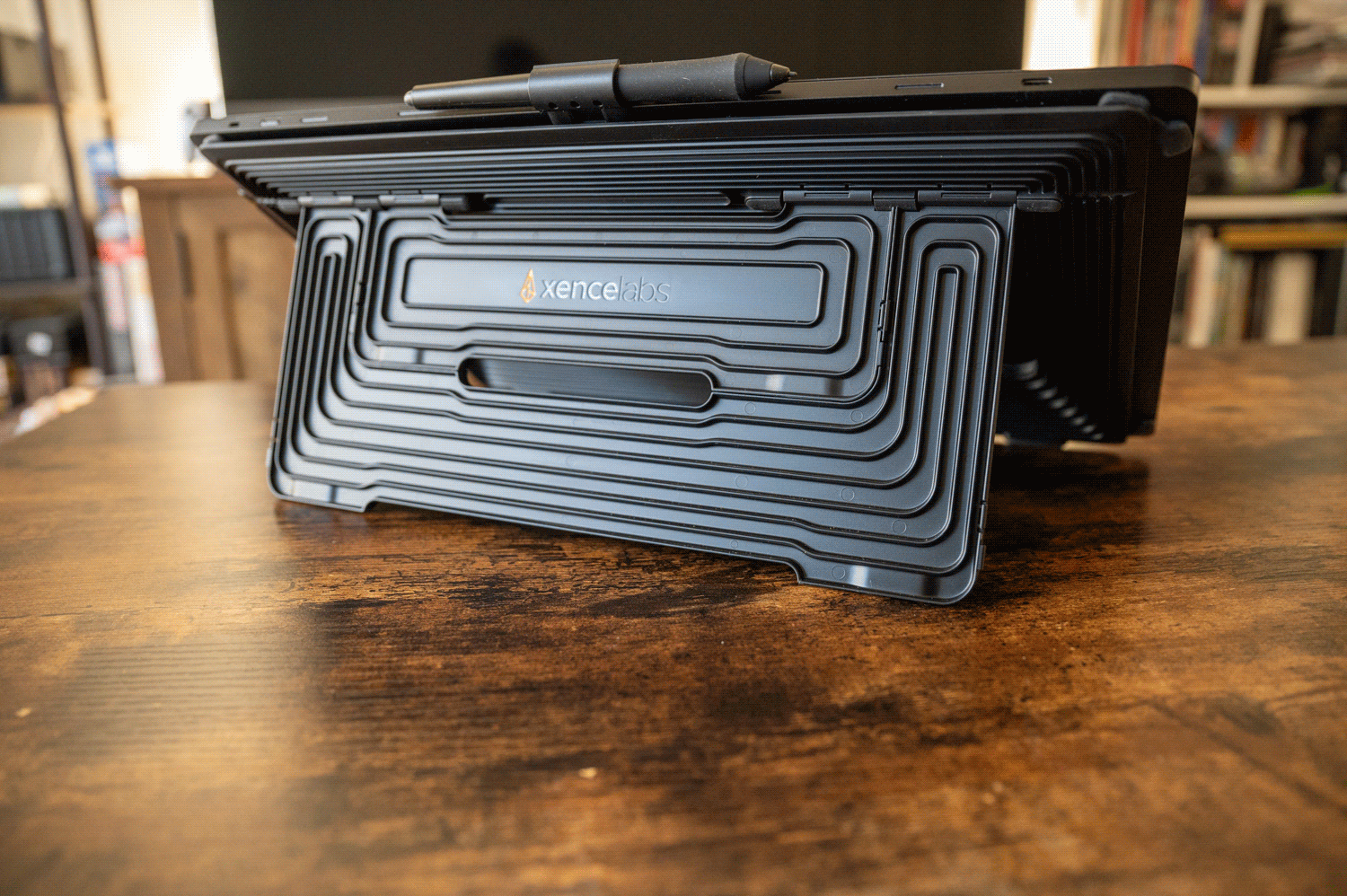 A graphics tablet is positioned on a wooden desk with its back facing the camera. A pen rests on top of the tablet. The back features a unique ridged design with a logo reading "XenceLabs." Bookshelves can be seen blurred in the background.