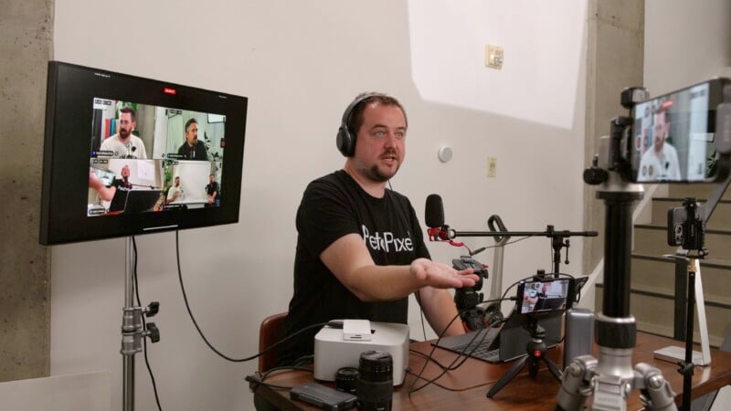A man wearing headphones sits at a table with podcasting equipment, including a microphone, camera, laptop, and mixer. He gestures while speaking. A monitor next to him displays multiple video feeds, and another camera on a tripod records him from the front.