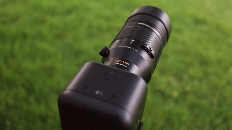 A black camera with a zoom lens attached, positioned and ready for a photograph. The settings on the lens indicate a focal range of 100-400mm. The background is a blurred green field, suggesting an outdoor setting.