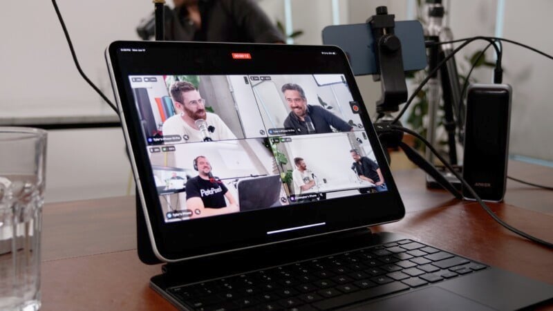 A tablet on a desk displays a video conference with five participants, each in their own window. A phone is mounted on a tripod beside the tablet. A glass of water and various cables are on the desk. A person is partially visible in the background.