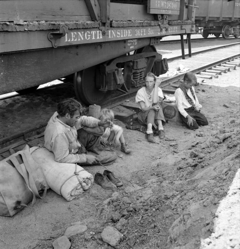 A black and white photo of four people sitting by the side of a train car on a dirt ground. Two adults and two children are seen, with the adults appearing weary. One child sits on an adult's lap, while the other child sits alone, resting their head on their hand. Bags and belongings are nearby.