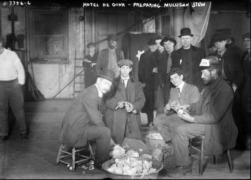 A group of men in early 20th-century attire sit and stand inside a large room, peeling potatoes and preparing Mulligan stew. The setting is “Hotel de Gink,” and there is a flag with a star visible in the background. The mood appears communal and industrious.