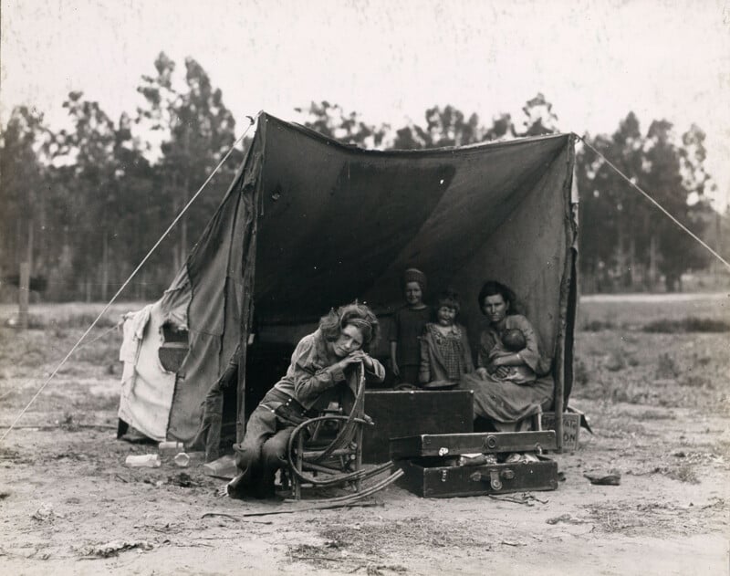 A black and white photo of a family at a makeshift campsite. A woman sits inside a tent holding a baby, with two children standing next to her. A boy leans on a box outside the tent. Trees are visible in the background. Suitcases and belongings are scattered.