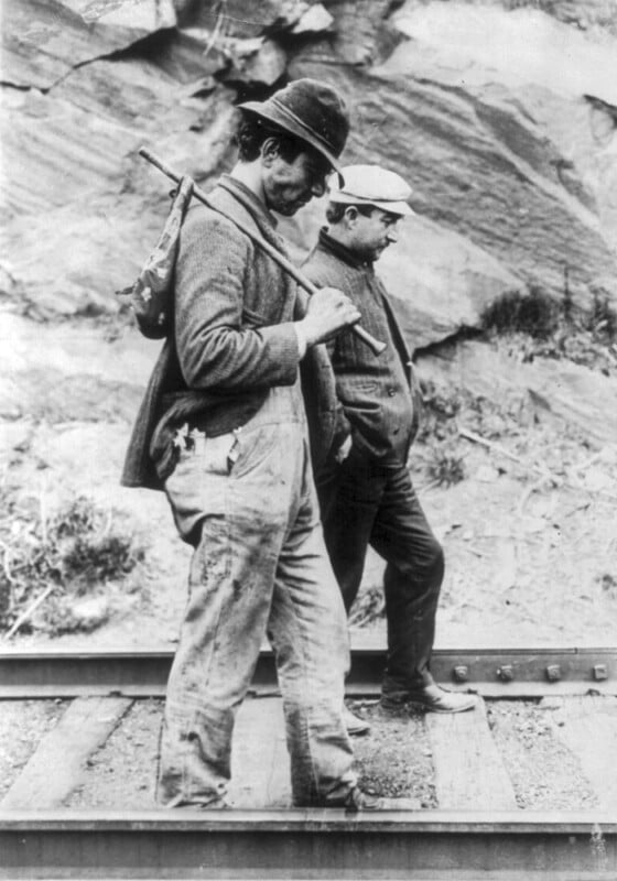 Black and white photo of two men walking side by side on a train track, with rocky terrain in the background. One man carries a bag over his shoulder with a stick, while the other looks down, walking with his hands in his pockets. Both wear hats and casual clothing.