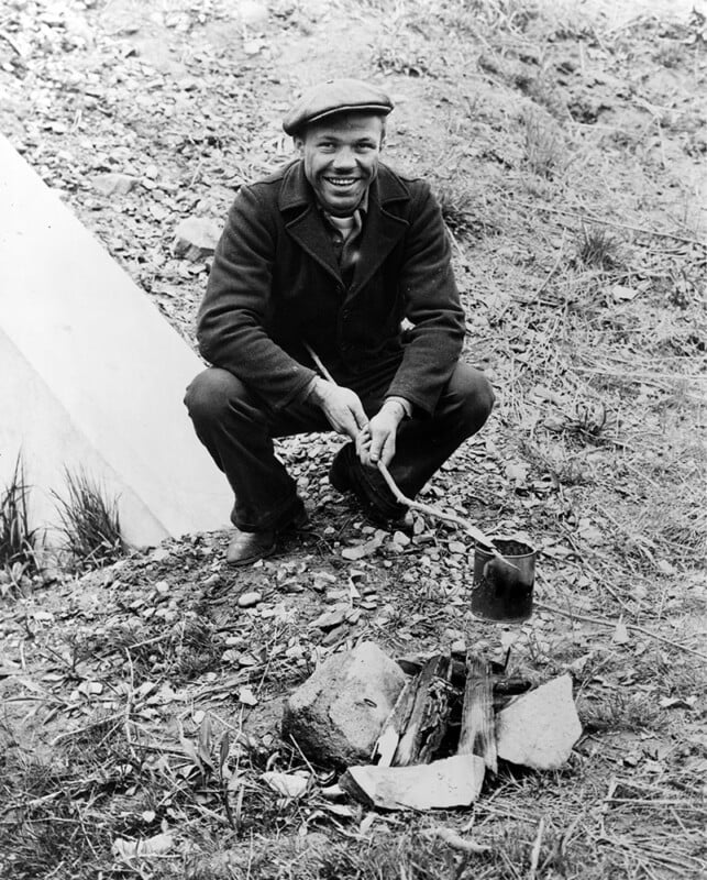 A man squats near a small campfire on a rocky patch of ground. He wears a cap, a dark coat, and pants, smiling broadly while stirring a pot over the fire with a stick. The surroundings are rustic with sparse vegetation and a slope in the background.