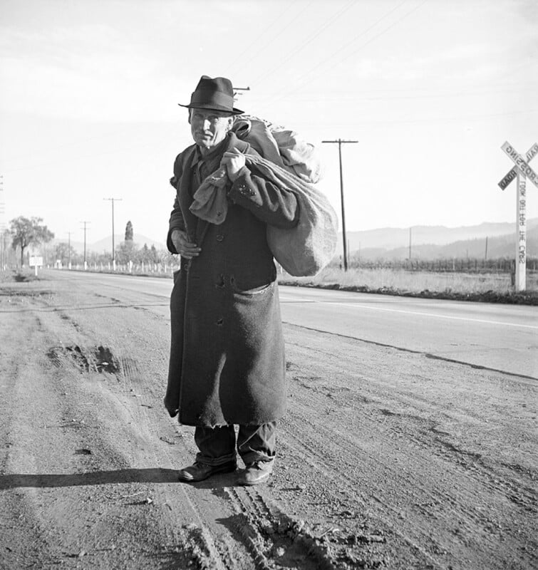 Black and white image of a man wearing a long coat and hat, standing on a dirt road, carrying a large sack over his shoulder. Utility poles and a railroad crossing sign are visible in the background, with fields and hills in the distance.