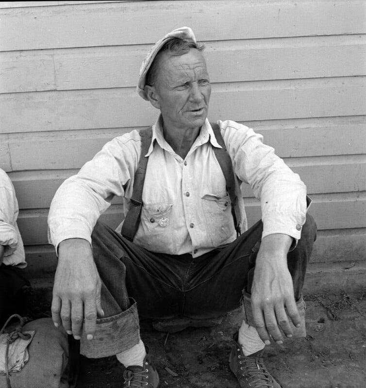 A man in a button-down shirt, rolled-up jeans, suspenders, and a hat sits on the ground against a wall. He looks off to the side with his hands resting on his knees. There is a bag partially visible next to him.