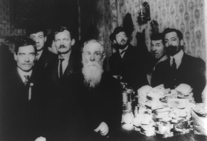A group of seven men, dressed in early 20th-century attire, stand in a dimly lit room. They are surrounded by stacks of bread and cans, perhaps representing a food distribution or gathering. The background features patterned wallpaper and a wall sconce.