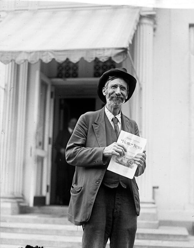 An elderly man with a beard and glasses stands outside a building holding a newspaper titled "HORO News." He is wearing a hat, jacket, and tie. A doorway with steps and columns is visible in the background.