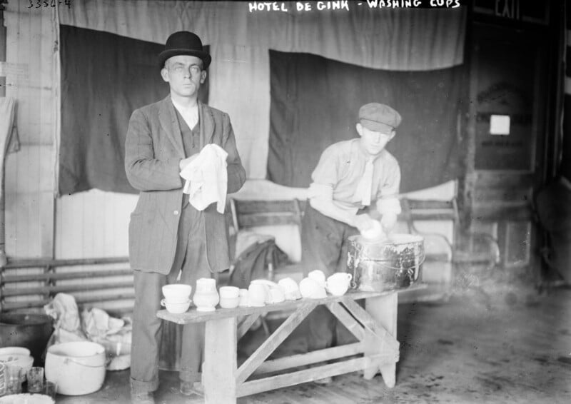 Two men are washing cups at the Hotel De Gink. One man, wearing a bowler hat and jacket, is drying a cup with a cloth. The other man, in a cap and shirt, is scrubbing cups in a washbasin on a wooden bench. Various cups and cleaning supplies are scattered around.