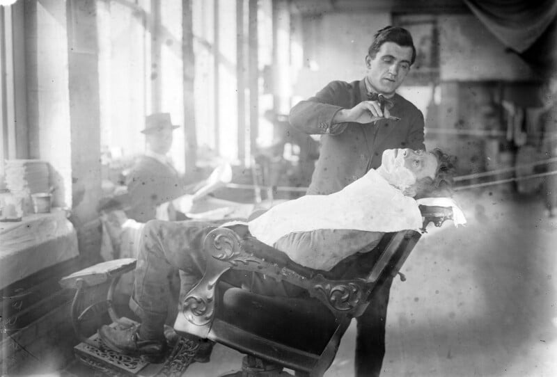 A barber is giving a shave to a customer who is reclining in a vintage barber chair in an early 20th-century barbershop. The barber is focused on the task while another person is seen in the background, seated and possibly reading a newspaper.