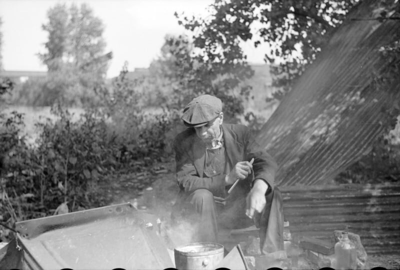 An elderly man in a flat cap and suit sits outdoors, stirring a pot over a smoky fire. He is surrounded by trees and foliage, with sunlight filtering through. A corrugated metal sheet leans against a tree in the background.