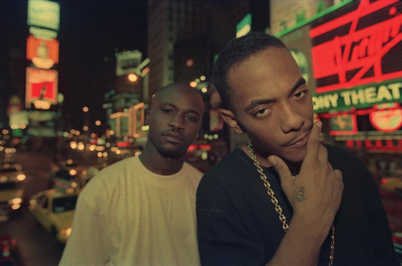 Two men are posing at night in a vibrant cityscape illuminated by neon signs and billboards. The man in the foreground has a thoughtful expression with his hand on his chin, wearing a sweater and a gold chain. The man in the background wears a light-colored shirt.