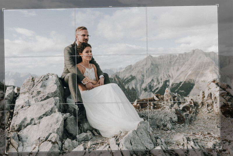 A bride and groom sit on a rocky mountain peak, with the groom sitting behind and slightly higher than the bride. Both wear formal wedding attire. The background features a mountain range under a partly cloudy sky. The scene conveys a sense of adventure and romance.