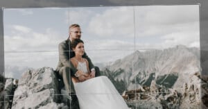 A bride and groom sit together on rocky terrain, with a mountainous landscape in the background. The groom is wearing a gray suit and glasses, while the bride is in a white wedding dress. They share an intimate and peaceful moment, surrounded by nature.