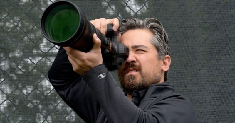A man with graying hair and a beard intently looks through a large zoom lens camera, aiming it upward. He wears a black jacket, and the background features a green mesh fence. The camera is pixelated, obscuring its make and model.