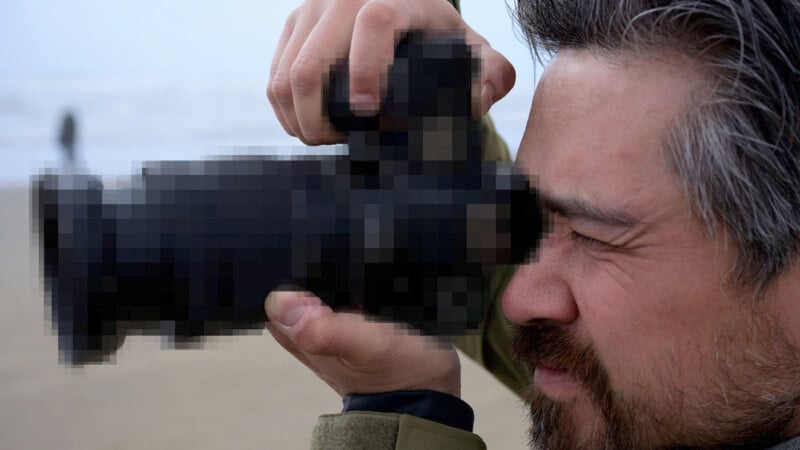 A man with gray-streaked hair is holding a camera up to his eye, focusing intently as he takes a photo. The background appears to be a beach with a figure blurred in the distance. The camera lens and viewfinder are pixelated.