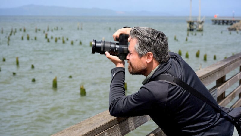 A person with gray hair and beard, wearing a black jacket, leans against a wooden railing to take a photo with a DSLR camera. They are overlooking a large body of water with wooden posts sticking out, and land is visible in the distance.