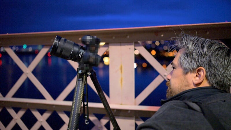 A person with gray hair, wearing a dark jacket, looks through a camera mounted on a tripod. The scene takes place at night on a bridge with blurred city lights in the background. The camera equipment is slightly pixelated.