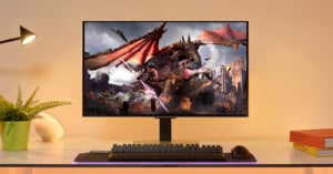 A monitor on a sleek white desk displays a vibrant fantasy scene featuring a dragon and warriors in combat. The setup includes a keyboard and mouse on a glowing mat. A small plant, books, and a desk lamp are also on the desk. The background is softly lit.