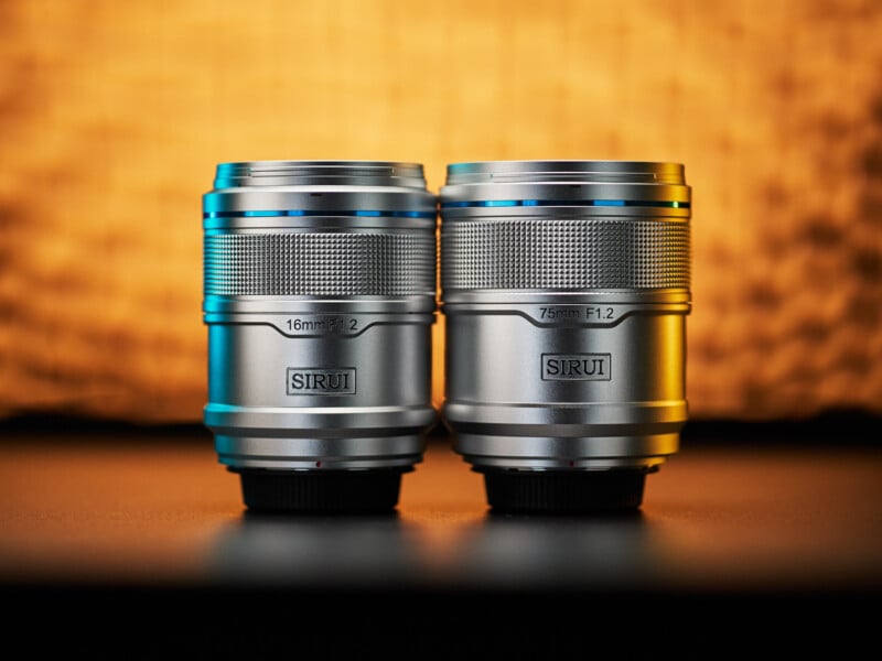 Two silver camera lenses, one labeled "16mm F1.2" and the other "75mm F1.2", stand upright on a dark surface against a warm, blurred background. Both lenses have the "SIRUI" brand logo visibly printed on them.
