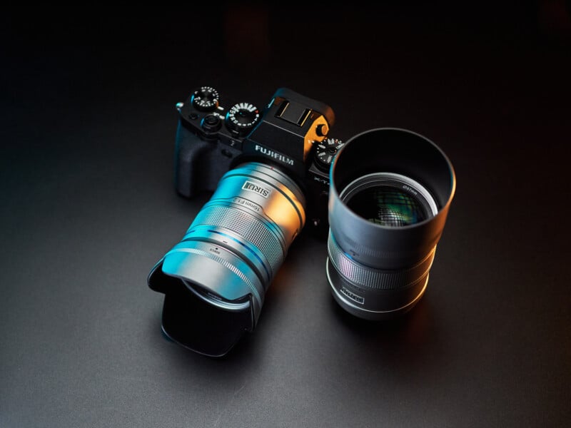 A Fujifilm camera with a mounted Zeiss lens sits on a black surface, accompanied by a second Zeiss lens. The setup is highlighted by moody lighting, with blue and orange tones reflecting off the camera and lenses.