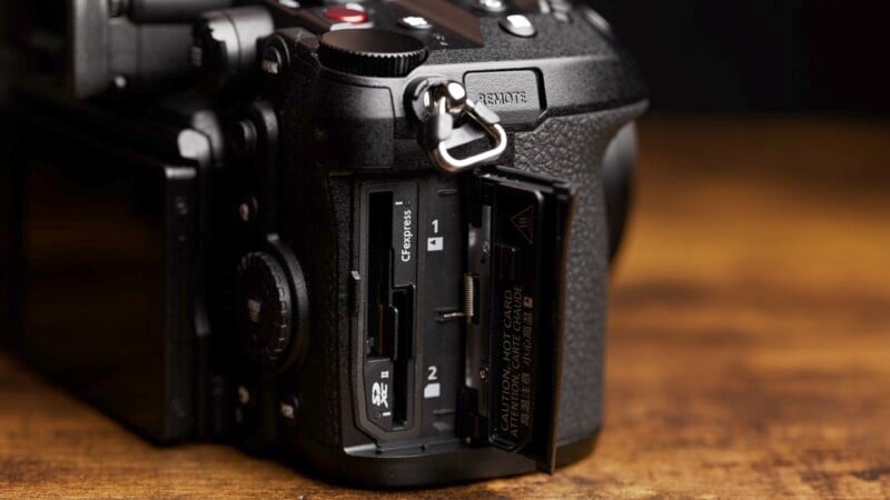 Close-up of an open memory card slot on a DSLR camera, positioned on a wooden surface. The slot contains compartments for a CFexpress Type B card and an SD card. The camera body is black and has a textured grip.