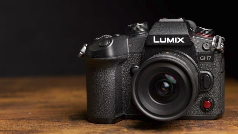 A black Lumix GH7 camera is positioned on a wooden surface against a dark background. The camera features a textured grip, various control dials, and a prominent lens with a red button on the front.