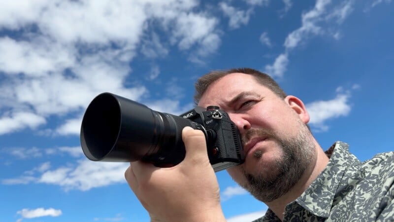 A man with a beard and wearing a patterned shirt holds a camera up to his eye, aiming it towards the sky. The sky behind him is bright blue with scattered clouds.