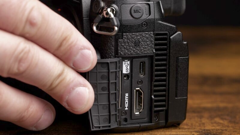 A close-up of a person's hand opening the side port cover of a DSLR camera. The revealed ports include a microphone jack, a USB-C port, and an HDMI port. The camera is black and the person has a light skin tone. The background is a wooden surface.