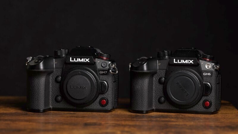 A close-up shot of two black Lumix cameras, model GH7 and GH6, placed side by side on a wooden surface against a dark background. Both cameras have red recording buttons and are without lenses, with Lumix lens caps attached on the front.