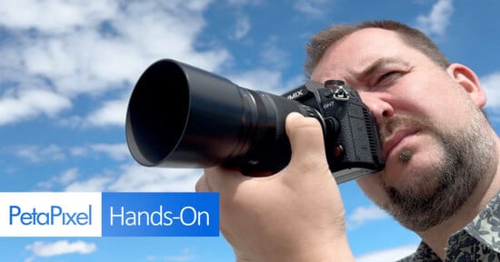A man is looking through the viewfinder of a Panasonic GH7 camera pointed towards the sky. The background features a vivid blue sky with scattered clouds. The text "PetaPixel Hands-On" is displayed in the lower-left corner of the image.