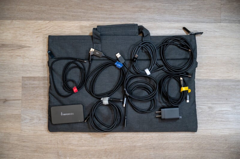 A variety of black cables and chargers with colored tags are neatly arranged on a grey bag with a handle. There is also a black power bank with the brand name "Anker" printed on it, placed on the left side of the bag. The background is a wooden surface.