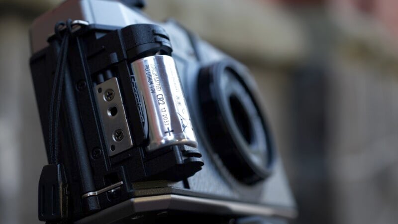 Close-up of a vintage camera with the battery compartment open, showing a silver battery inside. The camera appears to be mounted on a tripod, and the background is blurred, suggesting it was taken outdoors.