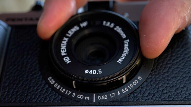 Close-up of a person's fingers adjusting the focus ring on a PENTAX camera. The lens has the specifications "HD PENTAX LENS HF 1:3.5 26mm Traditional" and the focus distance scale displays measurements in meters and feet ranging from 0.25 m to infinity.