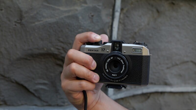 A hand holds an old-fashioned, black and silver Pentax 17 film camera against a textured, grey stone wall. The camera lens points directly at the viewer, and the details of the camera, including the focus ring and branding, are clearly visible.