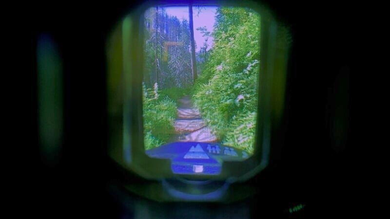 View through a night vision device showing a forest trail surrounded by dense greenery. The display includes directional and status indicators, with a clear path ahead.