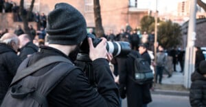 A person wearing a gray beanie and black jacket takes a photo with a large-lens camera in an urban setting. They have a black backpack, and several people are in the background, some blurred, suggesting a gathering or event.